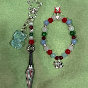Op de afbeelding: Een sleutelhanger met een stervormige sluiting, een doorschijnende lichtgroene vossenbedel en een metalen kunai-mes, naast een kralenarmband met rode, blauwe, witte en groene kralen, een zilveren vossenbedel en een rode torii-poortbedel.