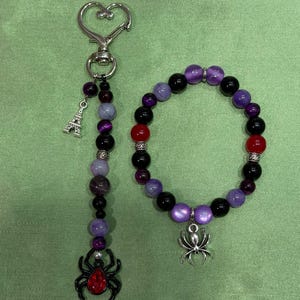 May include: A beaded keychain with a heart-shaped clasp, an Eiffel Tower charm, purple and black beads, and a spider charm with a red stone. Beside it is a beaded bracelet featuring purple, black, and red beads with a silver spider charm.