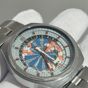 May include: A silver-tone stainless steel wristwatch with a round case and a metal link bracelet. The dial features a colorful map of the world with blue oceans and orange landmasses, a 24-hour scale, and the text EDOX printed near the bottom.