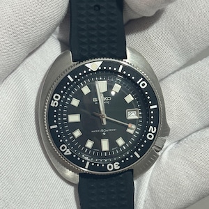 May include: A Seiko automatic wristwatch with a black dial, luminous hour markers, and a date window at the three o'clock position. The watch features a stainless steel cushion-shaped case, a black rotating bezel, and a textured black rubber strap.