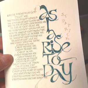May include: A calligraphic piece of art with gold and blue ink on a cream background. The text reads "May the strength of God pilot me... Christ in every ear that hears me." and "Rise to Day". The artist's name is "St. Patrick" in gold ink.