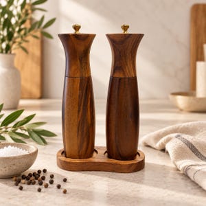 May include: A pair of tall, hourglass-shaped wooden salt and pepper grinders with dark brown wood grain patterns and small gold-toned metal knobs on top, resting together on a matching oval wooden base stand.