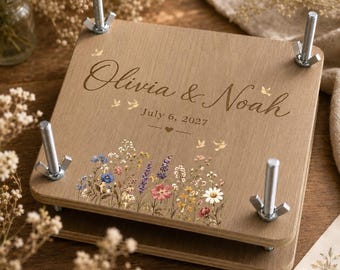 Könnte beinhalten: Eine quadratische Blumenpresse aus Holz mit vier Metallbolzen und Flügelmuttern an den Ecken. Die obere Platte ist mit dem Text Olivia & Noah, dem Datum 6. Juli 2027, einem kleinen Herz und einer bunten Illustration verschiedener Wildblumen graviert.