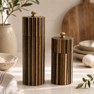 May include: Two cylindrical salt and pepper mills featuring a vertical striped pattern in alternating shades of dark brown, light brown, and black wood. Each mill is topped with a small, circular, metallic gold-toned adjustment knob.