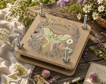 May include: A square wooden botanical press featuring four metal bolts with wing nuts at the corners. The top panel displays a printed illustration of a pale green luna moth surrounded by a colorful floral wreath and the text Emma Rose Botanical Press.