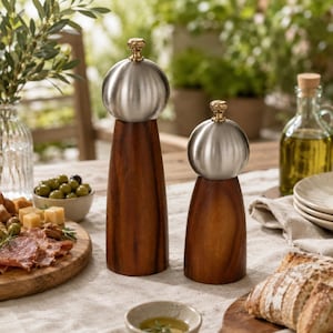 May include: Two salt and pepper grinders featuring dark brown wooden bodies with a tapered shape and brushed silver metal spherical tops. Each grinder is topped with a small gold-colored knob. The set stands on a light-colored fabric surface.