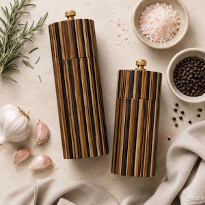 May include: Two cylindrical salt and pepper mills featuring a vertical striped pattern in dark brown and tan wood tones. Each mill is topped with a small, circular brass-colored metal knob for adjustment, providing a functional and sleek kitchen tool.