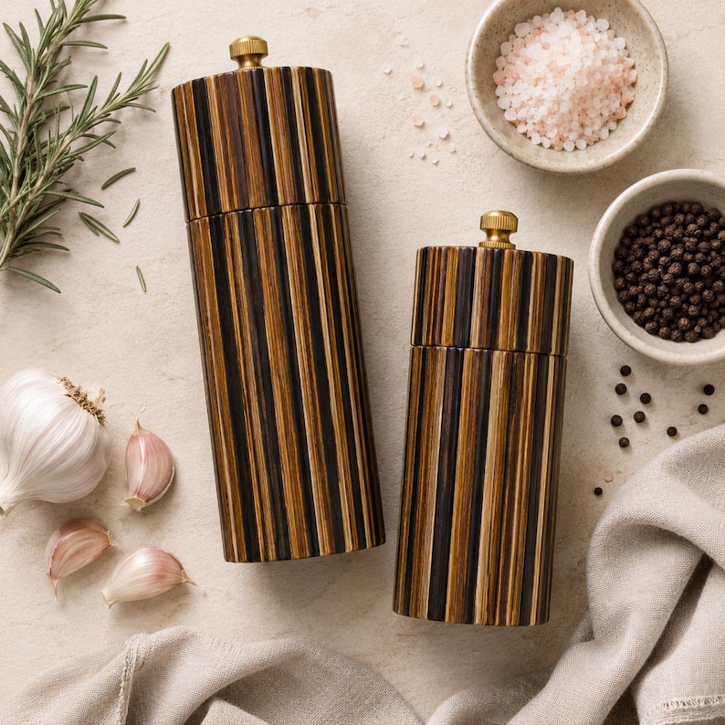 May include: Two cylindrical salt and pepper mills featuring a vertical striped pattern in dark brown and tan wood tones. Each mill is topped with a small, circular brass-colored metal knob for adjustment, providing a functional and sleek kitchen tool.