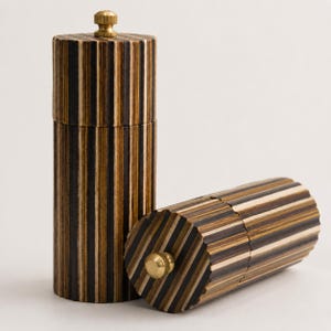 May include: Two cylindrical pepper mills featuring a vertical striped pattern of dark brown, light brown, and cream wood layers. Each mill has a fluted exterior surface and a small, circular, metallic gold-colored knob on the top end.