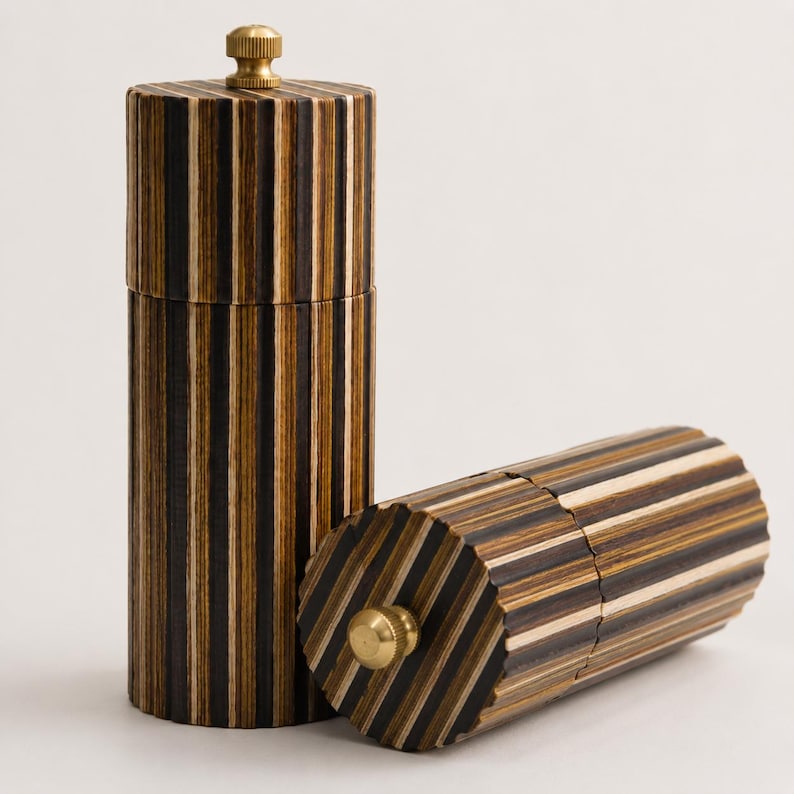 May include: Two cylindrical pepper mills featuring a vertical striped pattern of dark brown, light brown, and cream wood layers. Each mill has a fluted exterior surface and a small, circular, metallic gold-colored knob on the top end.