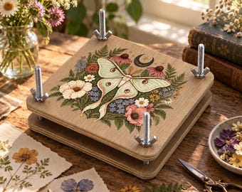 May include: A wooden flower press featuring a printed illustration of a pale green luna moth surrounded by colorful wildflowers and a crescent moon. The square press has four metal bolts with wing nuts at each corner to secure the wooden boards together.