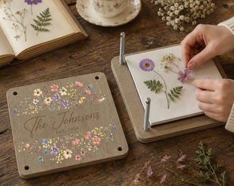 May include: A square wooden flower press with metal corner bolts, featuring a printed floral design and the text "The Johnsons EST. 2026". The press includes white paper sheets for drying pressed flowers, with a matching wooden top cover.