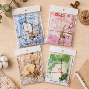 May include: Four clear plastic pouches containing assorted decorative paper materials in blue, pink, yellow, and green colour themes. Each pouch is tied with brown twine and features printed labels including The Blue Time, Pink Luxury Notes, Coffee Color Memory, and Green Listen To The Wind.