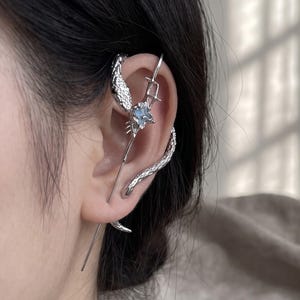 May include: A silver-toned ear cuff featuring a textured snake design that wraps around the ear cartilage. The piece includes a central square-cut iridescent stone and a long, slender metal pin that extends vertically along the ear structure.