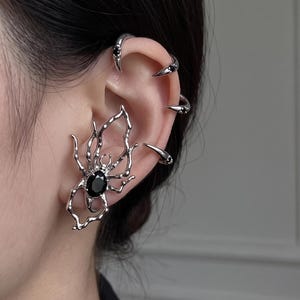May include: A silver-toned ear cuff shaped like a spider with a central black oval gemstone, accompanied by four smaller silver-toned ear cuffs along the upper ear cartilage, each featuring a tiny black stone at the pointed end.