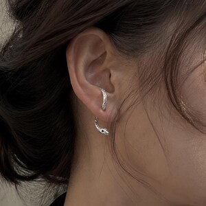 May include: A silver-toned ear cuff shaped like a slender snake with a textured, scaled surface. The jewellery wraps around the cartilage of the ear, with the head of the snake positioned at the top and the tail curving downward toward the earlobe.