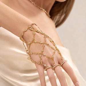 Könnte beinhalten: Ein goldfarbenes Sklavenarmband mit einem filigranen Kettenglied-Gitterdesign, das mit kleinen, klaren, rund geschliffenen Kristallen besetzt ist. Das Schmuckstück verbindet ein Armband mit drei einzelnen Fingerringen, die jeweils mit einer Reihe winziger, weißer Steine verziert sind.