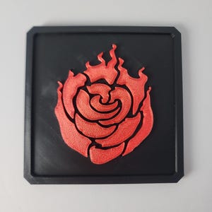 May include: A square black coaster featuring a raised, textured red rose design in the center. The rose petals are stylized with flame-like edges, creating a bold contrast against the flat, matte black background of the square base.