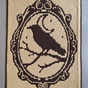 Puede incluir: Un tapiz rectangular de ganchillo que presenta la silueta de color marrón oscuro de un cuervo posado en una rama dentro de un marco ovalado ornamentado, sobre un fondo beige claro. El diseño incluye una luna creciente y estrellas.