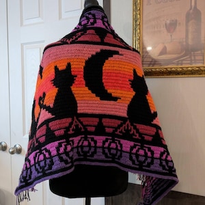 May include: A crocheted shawl featuring a gradient background of orange, pink, and purple yarn. The design includes black silhouettes of two cats, a crescent moon, and a row of pumpkins along the bottom edge, finished with fringe at the hem.