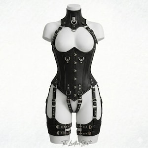 May include: A black leather-like corset featuring a high neck collar with a central metal ring, silver-toned hardware, and adjustable buckled straps. The garment includes integrated thigh garters and a front busk closure. The Leather Star logo is visible.