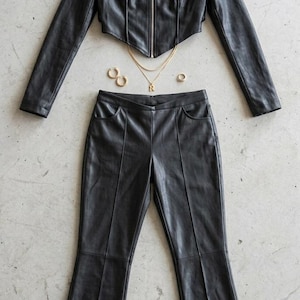 Black faux leather two-piece women’s outfit featuring a cropped zip-up jacket with stand collar and panel stitching, paired with matching high-waist flared pants, styled for edgy streetwear and modern fashion.