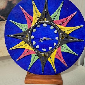 May include: A circular wall clock featuring a vibrant blue background with a starburst pattern created from black, yellow, pink, and light green string. The clock face includes small white stones as hour markers and ornate metal clock hands.