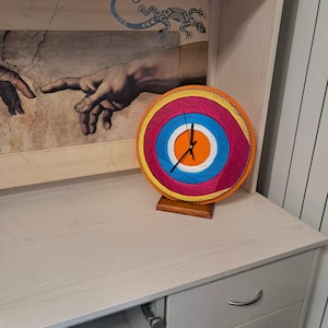 May include: A circular string art clock featuring concentric rings of orange, magenta, blue, and white thread. The clock has black hands and is mounted on a small wooden base. The design creates a vibrant, multi-colored pattern radiating from the center.