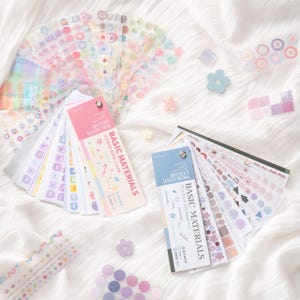 May include: Two fan-style sticker books labeled Basic Materials, one in Macaron Color and one in Morandi Color, featuring various adhesive shapes, patterns, and icons. Loose individual stickers in shapes like flowers and circles are scattered around.