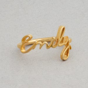 May include: A gold-toned ring featuring the name Emily written in a continuous cursive script. The metal band is integrated into the lettering, forming a single piece of jewelry with a smooth, polished finish and a distinct, flowing design.