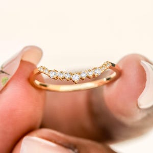 May include: A gold band ring featuring a curved, V-shaped design set with nine small, clear, round-cut stones arranged in a row along the top arc of the band. The metal has a polished, warm yellow finish, and the stones are evenly spaced.