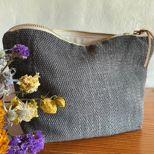 Könnte beinhalten: Eine dunkelgraue, gewebte Kosmetiktasche mit goldenem Reißverschluss und Lederlasche. Die Tasche ist geöffnet und zeigt ein beiges Innenfutter. Getrocknete Blumen in Lila, Gelb und Orange sind im Vordergrund.