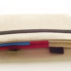 Puede incluir: Una cartera de tela beige con cierre de banda elástica negra. La cartera tiene un diseño en capas con inserciones de tela azul y roja visibles. Una pequeña etiqueta negra está adjunta al costado.