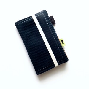 May include: A dark blue corduroy wallet with a white stripe down the center. The wallet has a black strap and a small green label with a black logo.