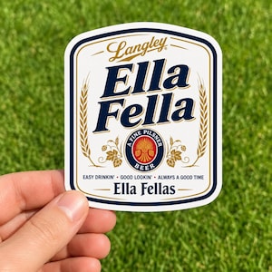Pode incluir: Um adesivo retangular branco com cantos arredondados apresentando o texto azul escuro Ella Fella, a palavra Langley em letra cursiva e um logotipo circular central com uma espiga de trigo. Inclui o texto Easy Drinkin' Good Lookin' Always A Good Time.