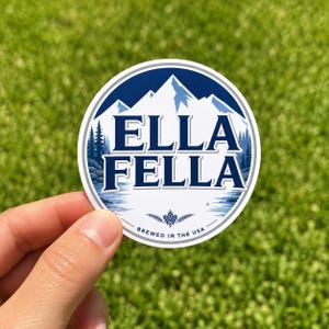 May include: A circular sticker featuring a dark blue and white mountain landscape with a forest of pine trees. The text ELLA FELLA is printed in large dark blue letters in the centre. Below, a small hop icon and the text BREWED IN THE USA appear.