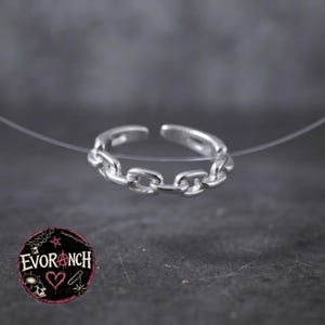May include: A silver-toned open ring featuring a continuous chain-link design. The ring is suspended by a thin, clear filament. In the lower left corner, a circular logo displays the text EVORANCH with a heart, star, and skull graphic.