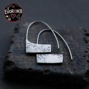 May include: A pair of silver-toned earrings featuring a curved wire hook and a rectangular pendant with a textured, hammered surface. The earrings are displayed with a circular logo in the corner that reads EVORANCH with a heart and star symbol.