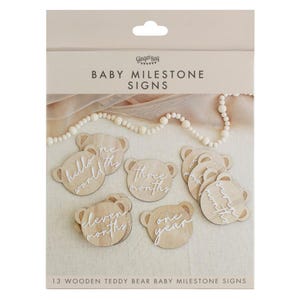 Op de afbeelding: Een set van 13 houten baby mijlpaalkaartjes in de vorm van teddybeerhoofdjes. Elke lichtbruine houten schijf is voorzien van witte schrijfletters die verschillende tijdsintervallen aangeven, zoals hello world, three months en one year.