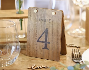 May include: A tent-style table number sign made of wood with a visible grain pattern. The sign features the word TABLE printed above the large numeral 4 in a dark font. Two small metallic fasteners secure the top of the two wooden panels together.