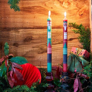 May include: Two tall, slender taper candles featuring a nutcracker soldier design. One candle is primarily green and the other is red, both with white, black, and gold painted details. Each candle is lit and sits in a dark, textured glass holder.