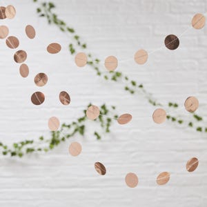 May include: A decorative garland featuring numerous metallic rose gold circular discs strung together on a thin white thread. The shiny, reflective paper circles are evenly spaced along the string, creating a cascading effect of round shapes.