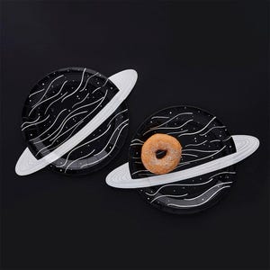 May include: Two round black paper plates shaped like planets with white ring attachments. The plates feature a white wavy line and dot pattern resembling outer space. One plate holds a single round golden-brown glazed donut in the center.