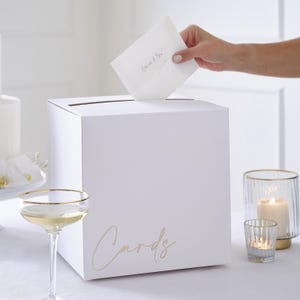 Könnte beinhalten: Eine quadratische weiße Kartenbox mit dem Wort Cards in goldener Schreibschrift auf der Vorderseite. Die Box hat einen horizontalen Schlitz an der Oberseite zum Einwerfen von Umschlägen. Das Design ist schlicht und für das Sammeln von Karten.