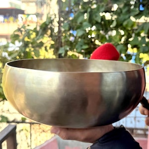 May include: A metallic gold-toned singing bowl with a smooth, polished exterior surface and a wide, open circular rim. A small mallet with a bright red felt head is positioned near the edge of the bowl, ready for use in producing resonant sound.