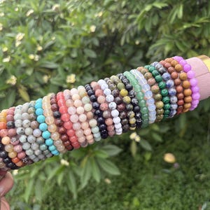 Könnte beinhalten: Eine Sammlung zahlreicher Perlenarmbänder, die in einer Reihe auf einem zylindrischen Präsentationsständer angeordnet sind. Die Armbänder weisen verschiedene Farben und Texturen auf, darunter Rosa, Blau, Grün, Braun, Schwarz, Weiß und Gelb.