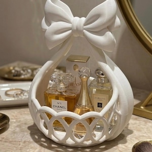 May include: A white 3D-printed basket with a large bow at the top and a lattice-patterned base. The basket holds three glass perfume bottles, including Chanel No. 5, Dior J'adore, and Jo Malone, which contain amber and clear liquids.