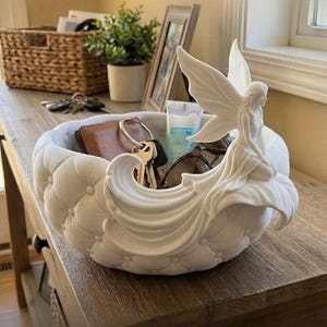 May include: A white ceramic bowl featuring a tufted, button-pressed exterior texture and a sculpted fairy figure perched on the rim. The bowl contains a brown leather wallet, a set of metal keys, a tube of lotion, and dark-rimmed eyeglasses.