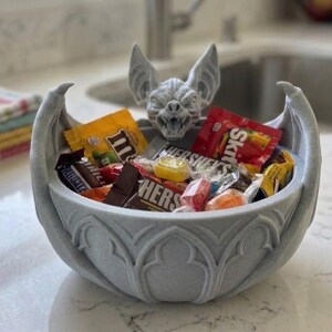 May include: A light gray bowl featuring a sculpted bat head at the center and bat wings extending along the sides. The bowl is filled with various colorful, individually wrapped candy pieces including M&Ms, Skittles, and Hershey's chocolate bars.