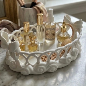 May include: A white oval decorative tray with a lattice-patterned side wall, featuring sculpted bow accents on the ends and a swirling base design. The tray holds several glass perfume bottles with gold caps and a small beige tassel accessory.